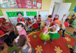 Grupa dzieci śpiewa piosenkę do mikrofonów, kilkoro dzieci naśladuje grę na gitarze z wykorzystaniem rekwizytu. Kilkoro dzieci tańczy.