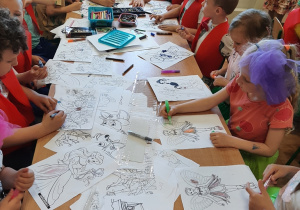 Dzieci siedzą przy podłużnym stole i kolorują kredkami obrazki z bohaterami z bajek.