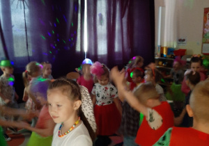 Dzieci w zaciemnionej sali przedszkolnej tańczą przy kolorowym, migającym oświetleniu.