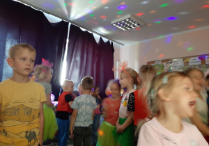Dzieci w zaciemnionej sali przedszkolnej tańczą przy kolorowym, migającym oświetleniu - inne ujęcie.