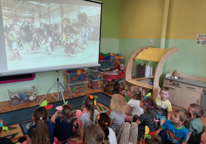 Dzieci siedzą w sali przedszkolnej na dywanie przed rozkładanym ekranem i oglądają pokaz indiańskich tańców.