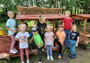 Grupa dzieci na placu zabaw pozuje do zdjęcia stojąc przy drewnianej ławce w kształcie lokomotywy.