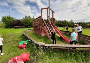Grupa dzieci bawi się na placu zabaw na drewnianym urządzeniu w kształcie statku, na którym jest zjeżdżalnia.