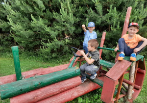 Trzech chłopców pozuje do zdjęcia siedząc na drewnianej zabawce - traktorze na placu zabaw.