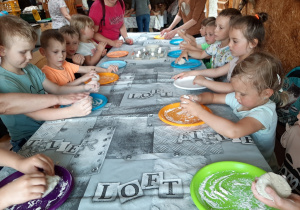 Dzieci siedzące przy podłużnym stole uczestniczą w warsztatach "Od ziarenka do bochenka", formują z ciasta chlebowego kształty pieczywa.