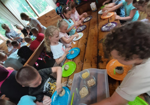 Dzieci siedzące przy podłużnym stole uczestniczą w warsztatach "Od ziarenka do bochenka", formują z ciasta chlebowego kształty pieczywa - kolejne ujęcie.