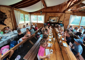 Dzieci siedzące na świetlicy przy podłużnych stołach spożywają zdrowe kanapki.