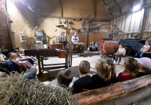 Dzieci siedzą w stodole słuchają opowieści animatora, oglądają stare sprzęty używane w gospodarstwach domowych.