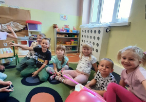 Dzieci siedzą na dywanie. Do oparć dwóch krzeseł odsuniętych od siebie przytwierdzono cienki sznurek, na który nawleczono kawałek słomki i przytwierdzono nadmuchany balon. Dzieci obserwują jaką siłę ma pęd powietrza wypływający z nadmuchanego balona.