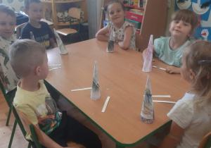 Dzieci siedzą przy stoliku i wykonują z papieru konstrukcję wiatraka.