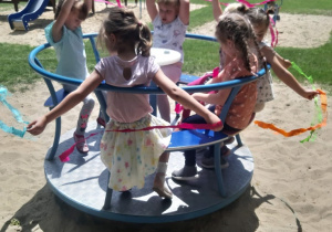 Dzieci na karuzeli na przedszkolnym placu zabaw sprawdzają pęd wiatru za pomocą pasków bibuły.