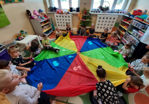 Dzieci z grupy 3,4,5 latków uczestniczą w zabawie z chustą animacyjną. segregują kolorowe koła z papieru kładąc je na odpowiednie kolory chusty.
