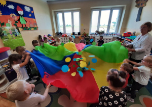 Dzieci z grupy 3,4,5 latków uczestniczą w zabawie z chustą animacyjną. Trzymają chustę stojąc w kole. Porusząjc nią podrzucają znajdujące się na niej koła z kolorowego papieru.