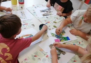 Grupa dzieci 5,6 letnich siedzi przy stoliku. wykonuje prace plastyczne tworząc różnokolorowe kropki za pomocą patyczków higienicznych maczanych w farbie.