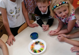 Grupa dzieci 5,6 letnich stoi przy stoliku. Wykonuje doświadczenie z użyciem kolorowych draży ułożonych na brzegu talerza i wody.