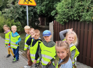 dzieci z grupy 5,6 latków na spacerze. Stoją na chodniku ubrane w kamizelki odblaskowe. W tle widać znak drogowy "uwaga dzieci".