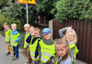 dzieci z grupy 5,6 latków na spacerze. Stoją na chodniku ubrane w kamizelki odblaskowe. W tle widać znak drogowy "uwaga dzieci".