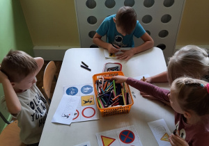 Kilkoro dzieci siedzi przy stoliku, ogląda plansze przedstawiające znaki drogowe, koloruje obrazki przedstawiające znaki drogowe.