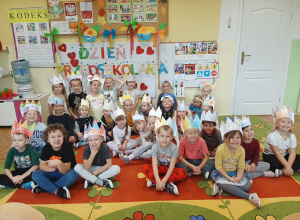 Dzieci z wartkowickiego przedszkola pozują w kolorowych papierowych koronach na tle gazetki tematycznej z okazji dnia przedszkolaka.