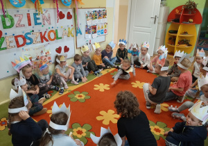Przedszkolaki w kolorowych papierowych koronach siedzą w kole na dywanie. W tle dekoracja z okazji dnia przedszkolaka.