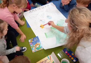 Dzieci w kręgu zapoznają się z kotem Amadeuszem poprzez kolorowanie plakatu z pomoca nauczyciela
