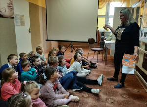 dzieci siedzą na dywanie w Gminnej Bibliotece Publicznej w Wartkowicach, słuchają ciekawostek na temat polskich naukowców przekazywanych przez Panią Bibliotekarkę.