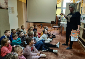 dzieci siedzą na dywanie w Gminnej Bibliotece Publicznej w Wartkowicach, słuchają ciekawostek na temat polskich naukowców przekazywanych przez Panią Bibliotekarkę.