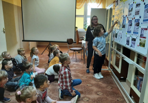 Dzieci siedzą na dywanie. chłopiec wraz z bibliotekarką stoją przy gazetce poświęconej polskim odkrywcom.