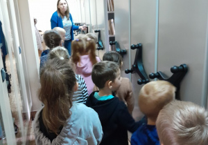 Dzieci stoją między regałami i słuchają pani bibliotekarki która opowiada o zasobach biblioteki
