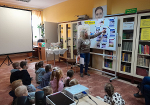 Pani bibliotekarka pokazuje dzieciom siedzącym na dywanie portrety polskich wynalazców