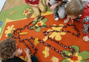 Chłopcy tworzą na dywanie sylwetkę kasztanowego ludzika. ozdabiają ją liśćmi i innym jesiennym materiałem przyrodniczym.
