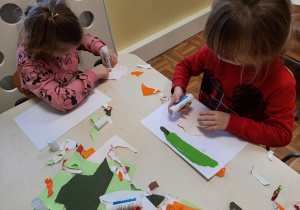 Dwie dziewczynki siedzą przy stole. Wydzierają z kolorowego papieru kształty warzyw i naklejają je na biały papier.