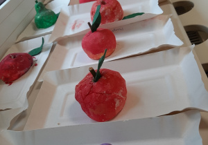 Wystawa prac przedstawiająca ulepione przez dzieci owoce z masy solnej. Owoce pomalowane są farbą, leżą na papierowych talerzach.