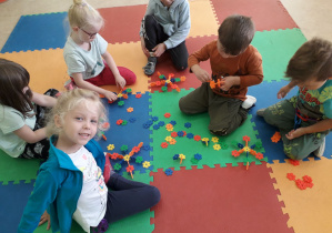 Grupa dzieci na dywanie bawi się klockami, konstruując różne stworki.