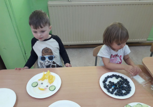Dzieci przy stolikach prezentują swoje warzywno-owocowe portrety.