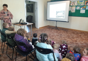 Dzieci w sali Muzeum siedzą na krzesłach, patrzą na prezentację multimedialną wyświetlaną na rozkładanym ekranie przez Panią Przewodnik.