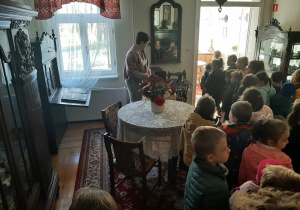 Dzieci zwiedzają Izbę Pamięci Konopnickiej, widać eksponaty: okrągły stolik, lustro, sekretarzyk i maszynę do szycia.