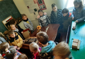 Dzieci zwiedzają Izbę Zbiorów Oświatowych, zasiadły i ustawiły się wokół dawnych ławek szkolnych, oglądają liczydło, piórnik z drewna.