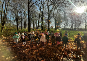 Dzieci w parku pozują podrzucając w górę zebrane liście, w tle drzewa i prześwitujące słońce.