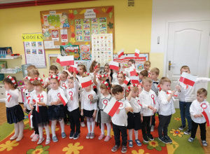 Dzieci na sali przedszkolnej pozują do fotografii ubrane na galowo. W rękach trzymają polskie flagi, do piersi maja przyczepione papierowe kotyliony.