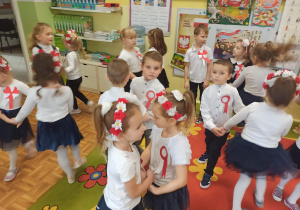 Dzieci ubrane w galowe stroje tańczą w parach. Dziewczynki mamą na głowach przepaski z białymi i czerwonymi kwiatami.