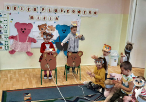 Dwoje dzieci z grupy 3,4,5 latków uczestniczy w zabawie - przeciąga sznurek przez otwór w papierowej sylwecie misia. Reszta grupy obserwuje siedząc na dywanie.