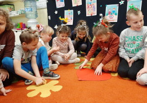 Grupa dzieci siedzi na dywanie, uczestniczą w andrzejkowej wróżbie. Dziewczynka nakłuwa szpilką papierowe czerwone serce z imionami chłopców.