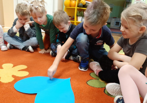 Grupa dzieci siedzi na dywanie, uczestniczą w andrzejkowej wróżbie. Chłopiec nakłuwa szpilką papierowe niebieskie serce z imionami dziewczynek.