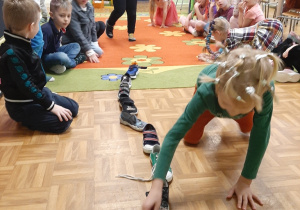 Dzieci siedzą na dywanie biorą udział we wróżbie "wędrujące buty". Chłopcy przestawiają buty.