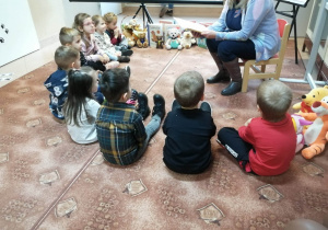 Dzieci siedzą w kole i słuchają opowiadania czytanego przez panią bibliotekarkę