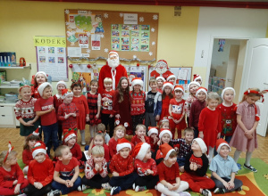 Dzieci z Mikołajem pozują do fotografii na tle zimowo - świątecznej dekoracji. Przedszkolaki ubrane są na czerwono, mają na głowie czapki Mikołaja.