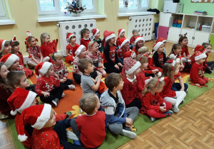 Dzieci siedzą na dywanie ubrane w czerwone stroje i czapki Mikołaja.