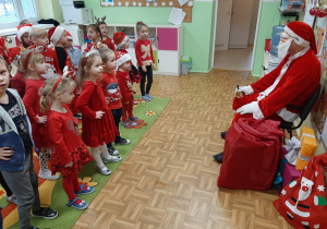 Dzieci recytują w grupie wiersz, którego słucha Mikołaj siedzący na przeciw nich na krześle, inne ujęcie.