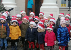 Grupa dzieci stoi przed wejściem do pałacu w Starym Gostkowie. Przedszkolaki mają na sobie świąteczne, mikołajowe czapki, śpiewają.